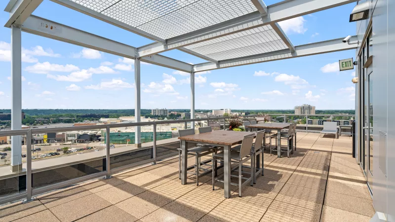 Bright rooftop patio with two dining tables and chairs under a metal pergola, city skyline visible under a blue sky.