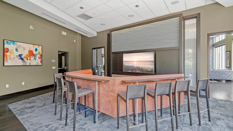 Spacious modern lounge with a curved, orange-brown bar, grey stools, a large TV, and colorful abstract art.
