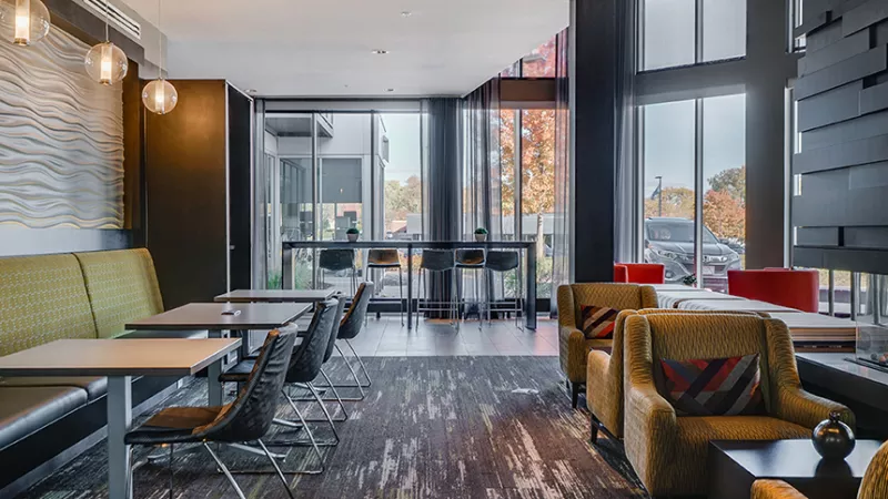 Modern lounge with yellow booth seating, patterned armchairs, high-top tables, and large windows.