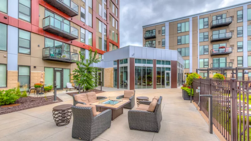Courtyard patio with modern lounge furniture, fire pit, and a glass-front clubhouse between apartment buildings.
