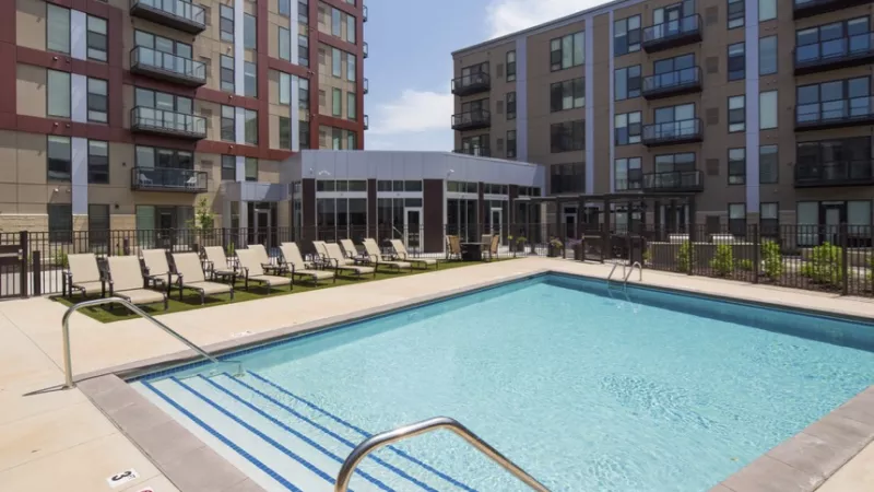 Outdoor swimming pool with lounge chairs, surrounded by modern apartment buildings and a clubhouse under a clear sky.