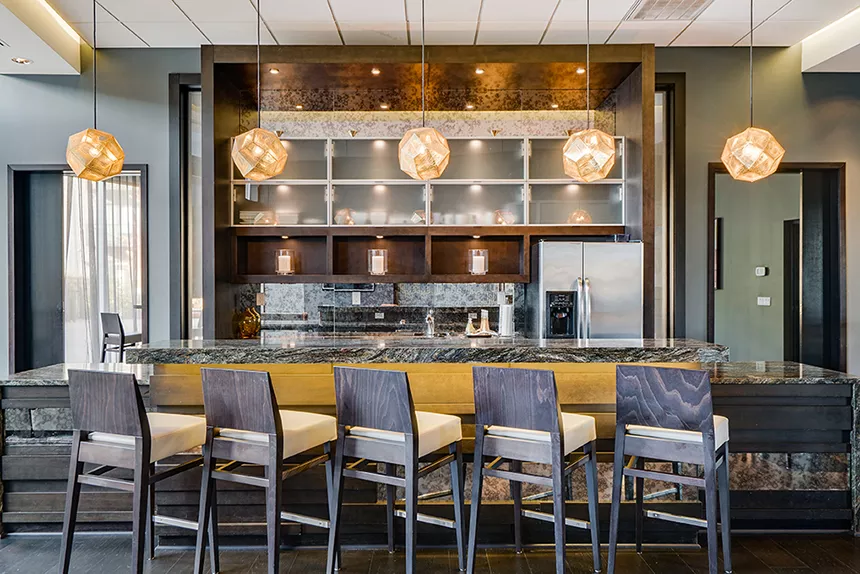 Modern bar with granite counter, six wooden stools, and warm spherical pendant lights over shelves with glassware and appliances.